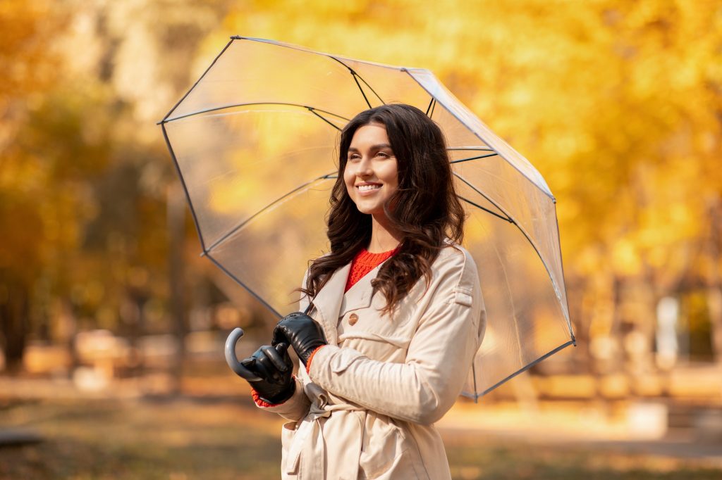 Autumn season concept. Fashionable young woman in stylish outfit holding umbrella and smiling at